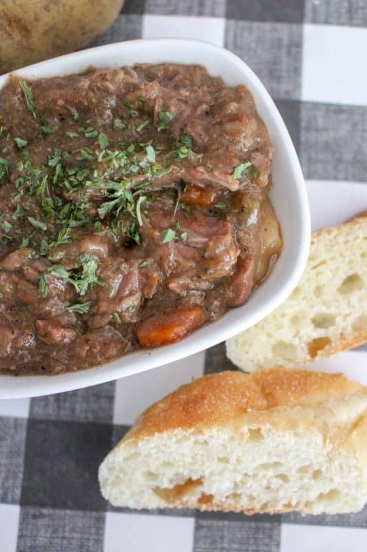 Slow cooker beef stew recipe in a white bowl with carrots, potatoes and bread in the background. 