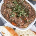 Slow cooker beef stew recipe in a white bowl with carrots, potatoes and bread in the background.