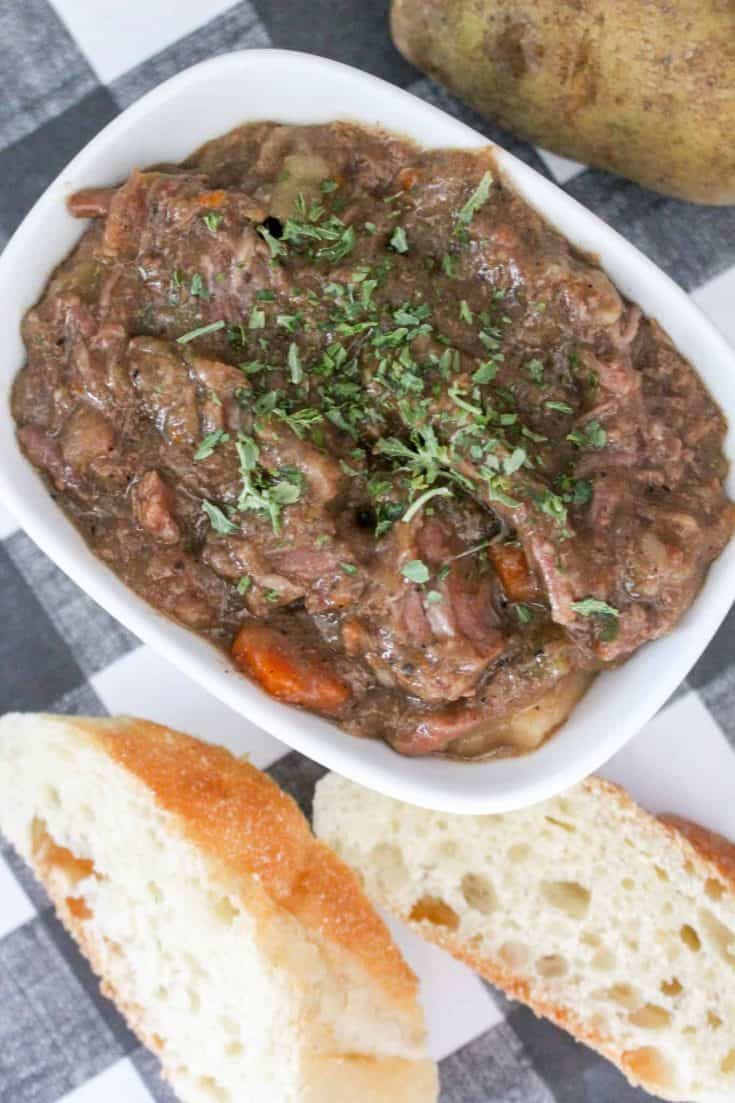 Slow cooker beef stew recipe in a white bowl with carrots, potatoes and bread in the background.