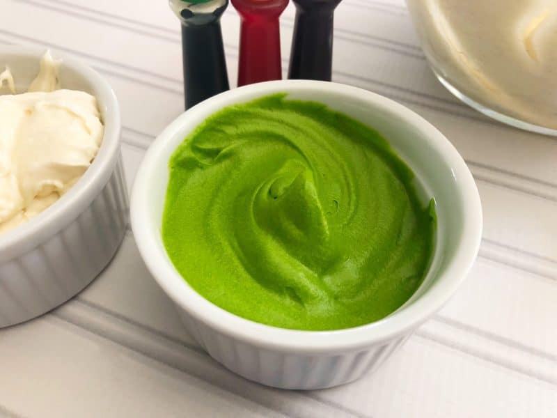 Bright green icing in a white bowl with 3 other gel colors behind it. 