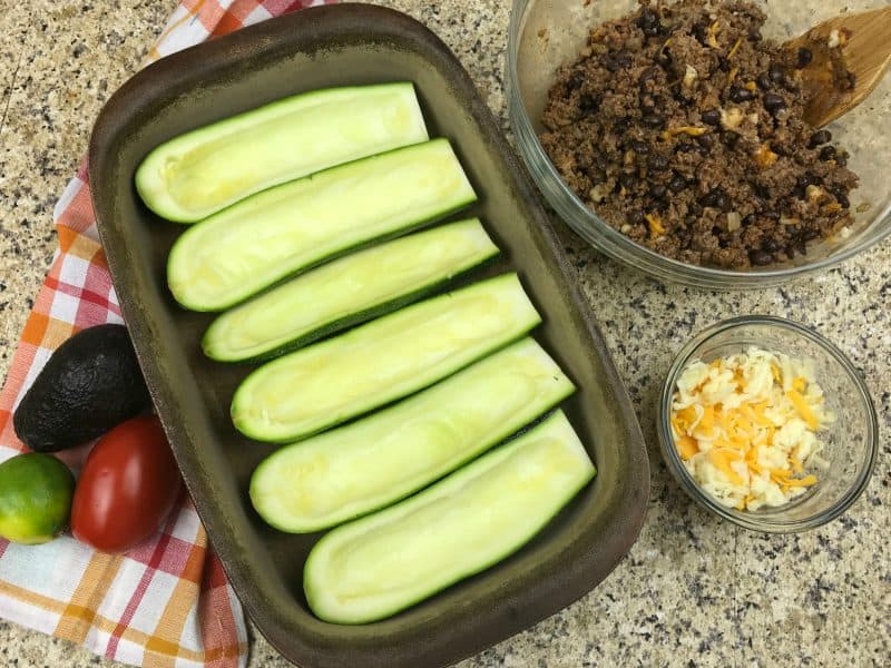 Hollowed out zucchini on a baking sheet with taco meat and cheese in bowls on the side. 